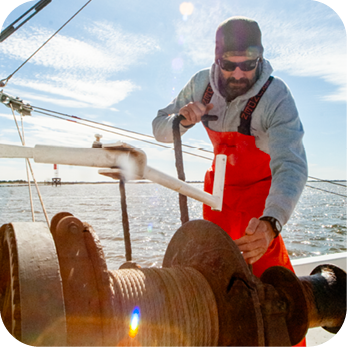  A commercial fisherman wearing a gray hoodie, red waterproof overalls, sunglasses, and a black beanie is working on a fishing boat. He is operating a large metal winch with one hand while gripping a lever with the other. The sun shines brightly, creating a lens flare effect. The background features open water with a distant structure visible on the horizon. The sky is mostly clear with a few wispy clouds.