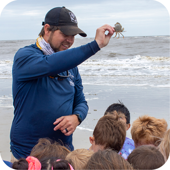 A male educator wearing a navy blue long-sleeve shirt, black cap, and patterned neck gaiter holds up a small crab while speaking to a group of children on a sandy beach. The children, with their backs to the camera, are attentively watching. The ocean waves roll onto the shore in the background under an overcast sky.