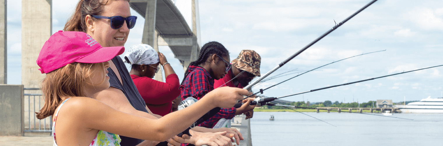 A diverse group of people, including children and adults, stand along a waterfront pier under a large bridge, fishing with rods over the railing. A young girl in the foreground wearing a bright pink hat and colorful tank top smiles as she holds a fishing rod with assistance from an adult woman in sunglasses. The background features calm water, a boat in the distance, and a partly cloudy sky.