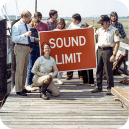 A group of people, including men and women dressed in casual and uniformed attire, stand on a wooden dock holding a large red sign that reads "SOUND LIMIT." One woman, wearing work clothes and boots, kneels in front of the sign while smiling at the camera. The background features a coastal marsh with tall grasses and calm water under a clear sky. Some individuals in the group are engaged in conversation, while others look at the sign or the camera.