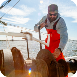  A commercial fisherman wearing a gray hoodie, red waterproof overalls, sunglasses, and a black beanie is working on a fishing boat. He is operating a large metal winch with one hand while gripping a lever with the other. The sun shines brightly, creating a lens flare effect. The background features open water with a distant structure visible on the horizon. The sky is mostly clear with a few wispy clouds.