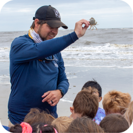 A male educator wearing a navy blue long-sleeve shirt, black cap, and patterned neck gaiter holds up a small crab while speaking to a group of children on a sandy beach. The children, with their backs to the camera, are attentively watching. The ocean waves roll onto the shore in the background under an overcast sky.