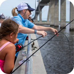 A young girl in a pink swimsuit and an adult in a blue shirt and white cap are fishing from a concrete pier. The adult is pointing and assisting the girl, who is holding a red and black fishing rod. Several other people are fishing in the background, and a large bridge stretches over the water. The water below is calm, and the sky is overcast.