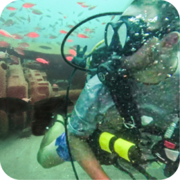 A scuba diver wearing a black diving mask, regulator, and a yellow oxygen tank explores an underwater artificial reef. The reef consists of large, hollow concrete structures resting on the ocean floor. Small red fish swim around the diver and the reef. Bubbles rise from the diver’s regulator, and the water has a greenish tint with particles suspended in it.